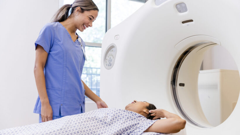 A mid adult woman talks to female radiographer before going through CT scan machine.