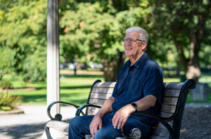 Older man sitting on a park bench outdoors, smiling, representing a patient whose life was saved by early heart screening.