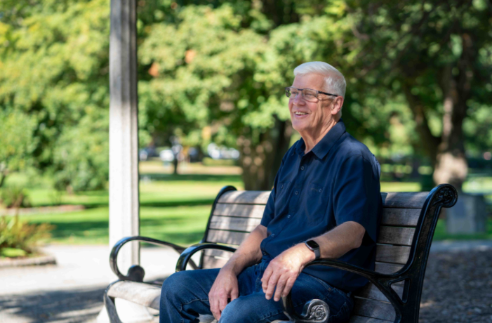 Older man sitting on a park bench outdoors, smiling, representing a patient whose life was saved by early heart screening.